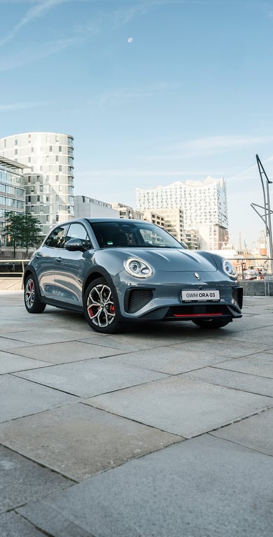 Der GWM ORA 03 GT in Meteorite Grey mit sportlichen Stoßfängern und Leichtmetallfelgen im GT Stil vor der Kulisse der Hamburger Elbphilharmonie